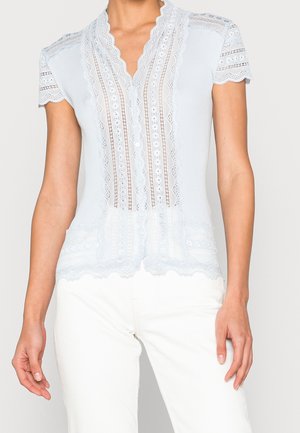 Biała bluzka z krótkim rękawem z koronkowymi zdobieniami, falistymi brzegami i guzikami z przodu, noszona z białymi spodniami przez stojącą osobę.