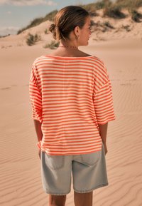 Striped orange and white oversized shirt with loose sleeves, paired with light denim shorts; background features sand dunes and sparse vegetation.