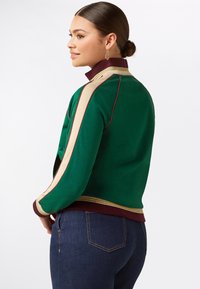 Vrouw met een bun in haar haar, gekleed in een groene jas met beige en bordeauxrode strepen en donkerblauwe jeans, kijkt opzij tegen een witte achtergrond.