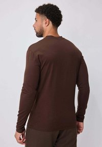 Chemise marron à manches longues en tissu doux, présentant un col rond et des manches ajustées, avec une texture lisse et sans motifs visibles.