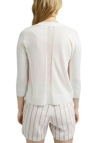 Vaaleanpunainen neulepaita, jossa on kolmivartiset hihat, takana on pystysuora avoin neuleyksityiskohta ja ribbaus. Yhdistetty raidallisiin shortseihin.