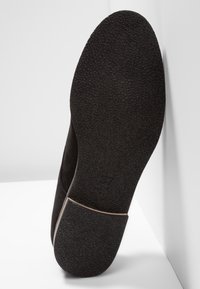 Chaussure en daim noir avec une semelle en caoutchouc texturée, dotée d'un talon arrondi et d'une étiquette de taille visible. Design épuré et simple avec des accents lisses.