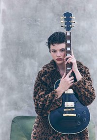Sort elektrisk guitar med guldtone, holdt op mod en tekstureret grå baggrund. Motivet bærer en leopardmønstrede pelsjakke.
