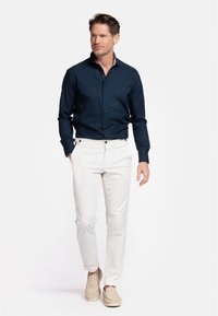 Chemise bleu marine à boutons avec un col à motifs, pantalon blanc et chaussures à enfiler beiges, mettant en avant une coupe ajustée et une texture de tissu lisse.