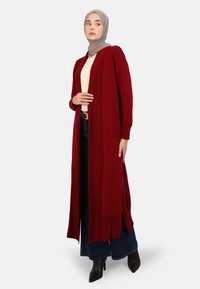 Long maroon cardigan with open front, ribbed texture, and long sleeves. Paired with a light top and wide-leg denim, finished with pointed heels.
