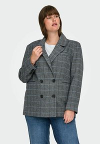 Femme portant un blazer à carreaux gris à double boutonnage sur un haut blanc et un jean bleu, regardant à sa gauche avec une main près du col.