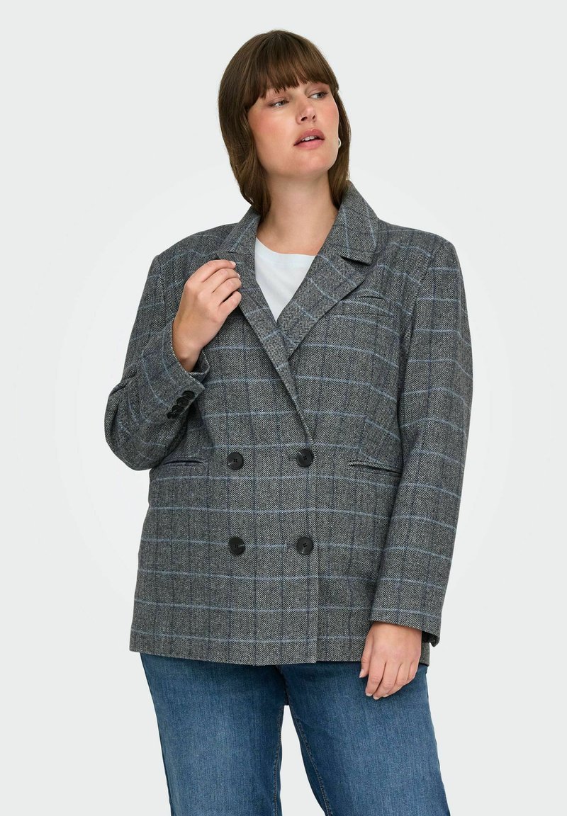 Femme portant un blazer à carreaux gris à double boutonnage sur un haut blanc et un jean bleu, regardant à sa gauche avec une main près du col.