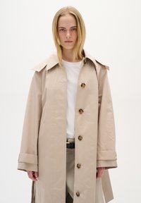 Béžový trench coat s velkým límcem, zapínáním na knoflíky a ohrnutými manžetami. Nosený přes bílou košili s hladkým, strukturovaným materiálem.