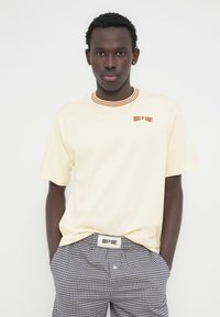 Ανοιχτό μπεζ κοντομάνικο μπλουζάκι με στρογγυλή, ριγωτή λαιμόκοψη και το κέντημα «HOUSE OF SUNNY» σε πορτοκαλί χρώμα. Συνοδεύεται από καρό σορτς.