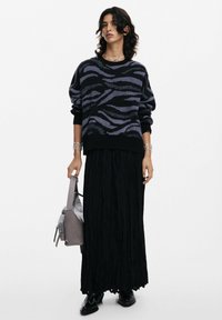 Maglione a righe nere e viola con una vestibilità rilassata, abbinato a una gonna lunga nera fluida. Tiene in mano una borsa grigia, con una texture liscia.