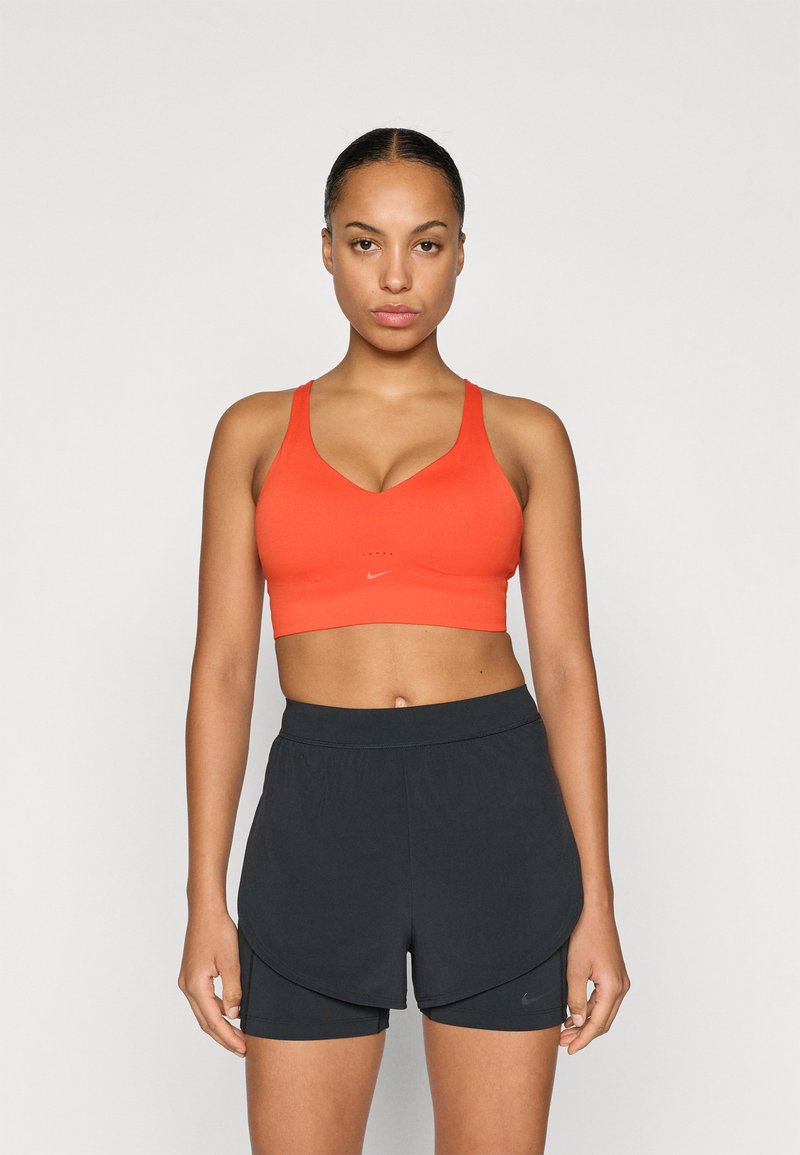 Oranje sportbeha met een V-hals en dikke banden, gecombineerd met zwarte losse shorts met een binnenlaag. Nike-logo zichtbaar.