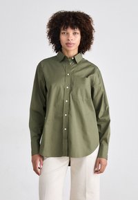 Olivengrønn skjorte med knapper laget av lett materiale, med brystlomme, lange ermer, og en buet kant. Har synlige hvite knapper.