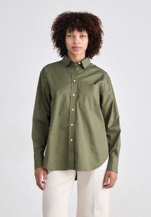 Olivengrønn skjorte med knapper laget av lett materiale, med brystlomme, lange ermer, og en buet kant. Har synlige hvite knapper.