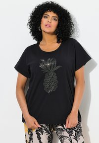 Musta lyhytnokkainen t-paita, jossa on hopeinen ananasgrafiikka, joka näyttää teksturoidulta. Yhdistetty kevyisiin, kuvioituihin vaaleisiin housuihin.