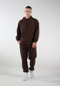 Brauner Kapuzenpullover mit Fronttasche, kombiniert mit passenden braunen Jogginghosen und weißen Turnschuhen. Schlichtes Design und weiche Textur.