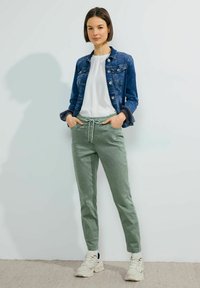 Chaqueta de mezclilla azul, camiseta blanca, pantalones verdes con cordón y zapatillas de color claro. Diseño sencillo con bolsillos y un corte relajado.