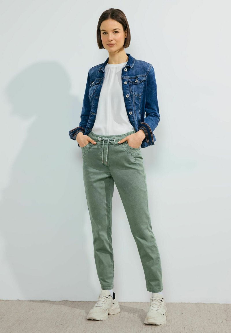 Chaqueta de mezclilla azul, camiseta blanca, pantalones verdes con cordón y zapatillas de color claro. Diseño sencillo con bolsillos y un corte relajado.