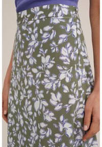 Kukkakuvioinen hame oliivinvihreästä kankaasta, jossa on valkoisia ja violetteja lehtikuvioita. Lähikuva näyttää tekstuurin ja sileän muotoilun sekä sivusauman.