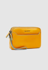 Gele leren pouch met een gladde textuur, blauwe accenten, ritssluiting en een polsbandje. Voorzien van een voorvak voor extra opbergruimte.