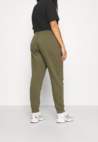 Persoon in olijfgroene joggerbroek, zwart T-shirt, witte sokken en witte sneakers, staand tegen een effen witte achtergrond.