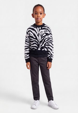 Barn stodende iført en sort og hvid zebra mønstret sweater med logo, mørke bukser og hvide sneakers på en ensartet lys baggrund.