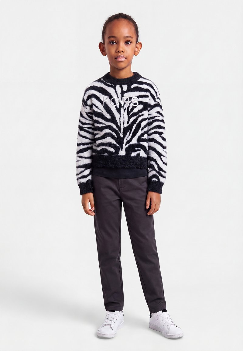Enfant debout portant un pull à motif zèbre noir et blanc avec un logo, un pantalon foncé et des baskets blanches sur un fond clair uni.