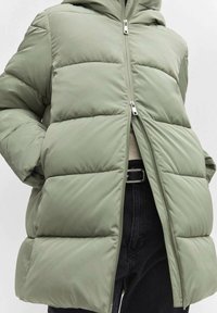 Hellgrüne Steppjacke mit Kapuze, gesteppten Abschnitten, Reißverschluss vorne und zwei Seitentaschen, getragen über einer schwarzen Hose.