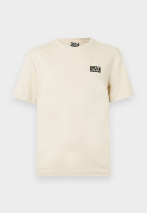 T-shirt en coton couleur crème avec des manches courtes, col rond et un petit patch logo noir EA7 sur la poitrine. Texture lisse, coupe décontractée.
