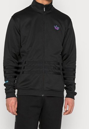 Homme portant une veste noire zippée avec un logo violet et bleu sur la poitrine et trois fines bandes horizontales noires sur les manches et à la taille.