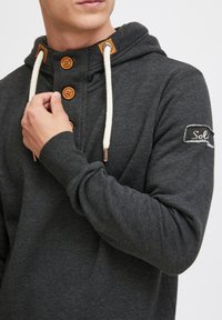 Sudadera gris oscuro con botones de madera, cordones blancos y acentos de cuero. Incluye un parche con la palabra "Sol" en la manga.