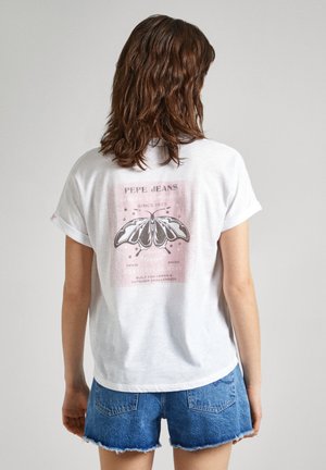 Camiseta blanca de algodón con un estampado gráfico de una mariposa y texto en rosa y negro en la parte posterior, con mangas cortas enrolladas, combinada con pantalones cortos de mezclilla.