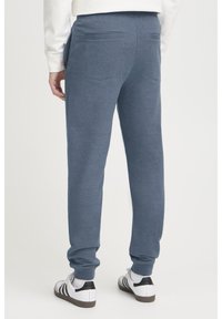 Pantalones de chándal grises con puños elásticos, hechos de una suave mezcla de algodón, con dos bolsillos traseros y un ajuste holgado. Zapatillas negras y blancas puestas.
