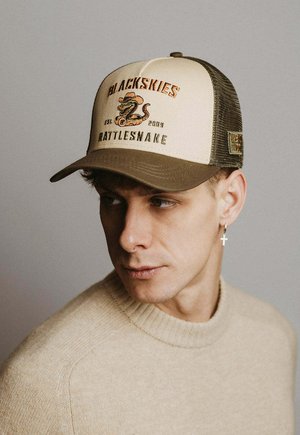 Mladenič, ki nosi bež pulover in bež-rjavo kapo z logotipom klopotca in besedilom "Blackskies Rattlesnake," gleda levo.