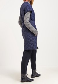 Marinblå quiltad väst med korta ärmar, bältesmidja och grå stickad tröja under. Svarta ankelskor och mörka byxor fulländar stilen.