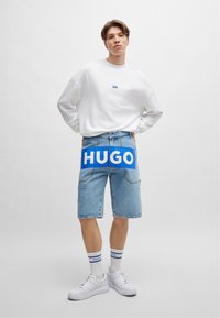 Sweatshirt en coton blanc avec un petit logo, shorts en denim bleu avec un grand graphique "HUGO", baskets blanches et chaussettes rayées.