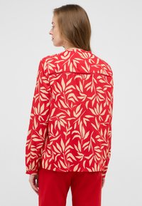 Blouse rouge avec un motif de feuilles en beige clair. Manches longues, coupe décontractée et un détail de empiècement étroit au dos. Texture du tissu lisse.