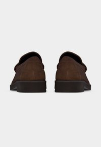 Καφέ σουετ παπούτσια χωρίς κορδόνια, εμφανιζόμενα από πίσω, με χαμηλές μαύρες λαστιχένιες σόλες, παρουσιάζονται σε απλό λευκό φόντο.