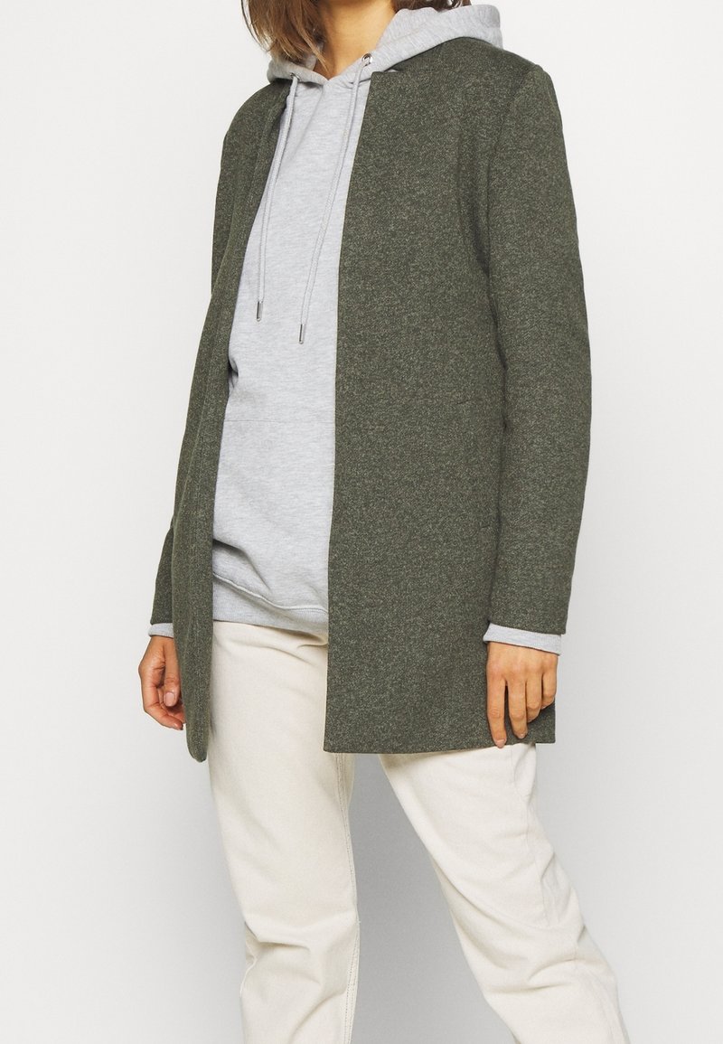 Personne portant un sweat à capuche gris sous un cardigan ouvert vert foncé avec un pantalon crème, debout devant un fond blanc.