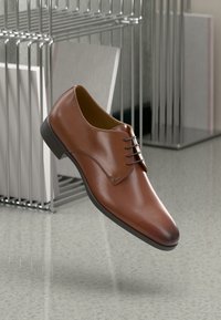 Brown leather dress shoe with a smooth finish, pointed toe, and three eyelets for laces. Black sole, minimal design, elevated position.