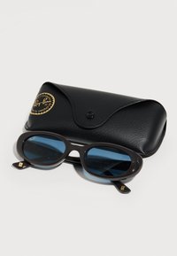 Γυαλιά ηλίου Ray-Ban με σκούρους μπλε φακούς και λεία μαύρη πλαστική κορνίζα, συνοδευόμενα από μια μαύρη υφασμάτινη θήκη με χρυσό λογότυπο.