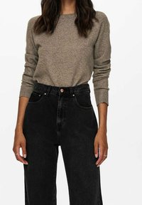 Beige gestrickter Pullover mit langen Ärmeln, kombiniert mit hoch geschnittenen schwarzen Jeans. Die Jeans verfügen über einen Knopfverschluss und Fronttaschen.