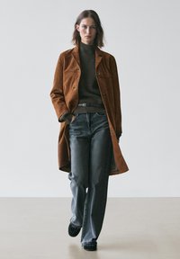 Bruine suède lange jas over een donkergeribbelde trui, gecombineerd met brede blauwe jeans en zwarte instapschoenen. Simpele, strakke lijnen en neutrale tinten.