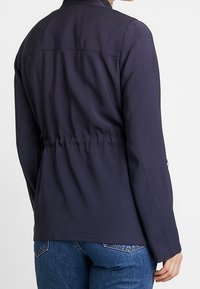 Giacca blu navy con vita cintrata, maniche lunghe e colletto alto. Presenta un tessuto strutturato e liscio con dettagli di cucitura.