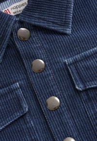 Niebieska koszula z materiału sztruksowego z metalowymi zatrzaskami i dwiema przednimi kieszeniami na klapie, ukazująca strukturę tkaniny oraz detal kołnierza.
