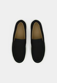 Μαύρα παπούτσια τύπου slip-on με ύφασμα στο πάνω μέρος, λευκή σόλα από καουτσούκ και λεία επιφάνεια. Διαθέτουν ελάχιστη ραφή και χωρίς ορατά μεταλλικά στοιχεία.