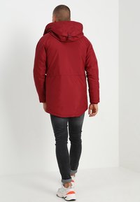 Chaqueta roja con capucha y aislamiento, con cintura fruncida, vista por detrás. Presenta una textura suave y puños elásticos, combinados con vaqueros oscuros.