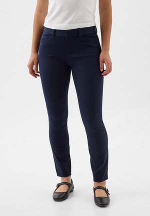 Mujer vistiendo pantalones ajustados azul marino, top blanco sin mangas y zapatos Mary Jane negros, de pie sobre un fondo blanco liso.