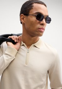 Beige teksturert langermet polo med glidelås krage. Modellen holder en svart veske over den ene skulderen og har på mørke solbriller.