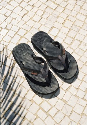 Svarte teksturerte flip-flops med grå stropper og oransje logoer på hvit flislagt gulv med skygge av et palmeblad.
