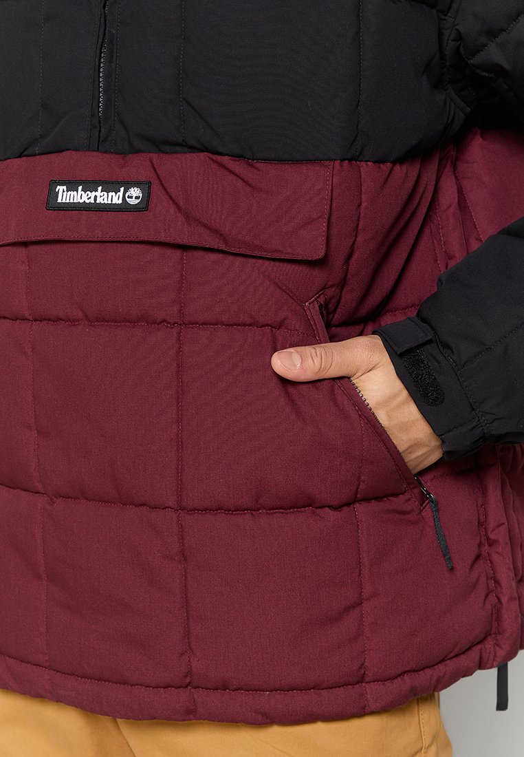 Osoba w bordowej i czarnej pikowanej kurtce Timberland, z ręką w bocznej kieszeni na zamek, w beżowych spodniach widocznych poniżej kurtki.